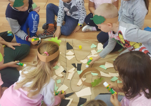 Grupa dzieci ozdabia wyciętymi elementami szablon drzewa narysowany na dużym arkuszu papieru pakowego.